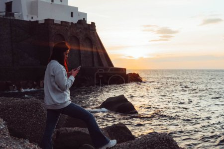 Foto de Woman looking at the phone in front of the sea at sunset - Imagen libre de derechos