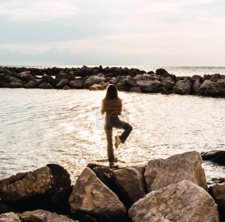 Foto de Woman on stones in front of the sea at sunset - Imagen libre de derechos