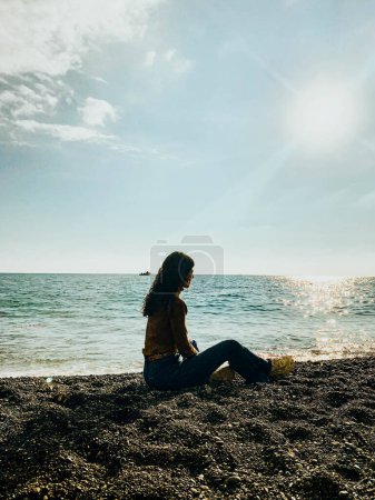 Foto de Woman sitting on black stone beach facing the sea looking at the sun - Imagen libre de derechos