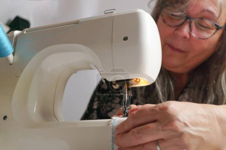 Foto de Older woman with white hair out of focus sewing a white fabric on a sewing machine - Imagen libre de derechos