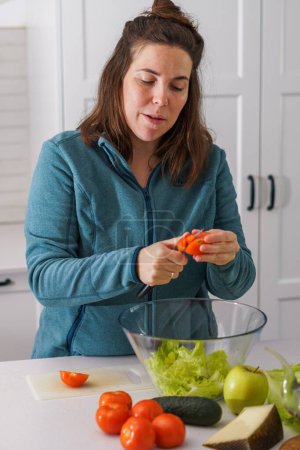 Téléchargez les photos : Beautiful young long-haired brunette girl cutting fresh tomatoes to prepare a salad in the kitchen - en image libre de droit