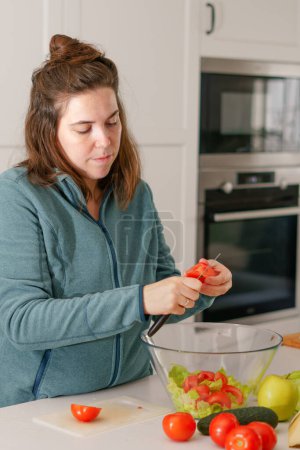 Foto de Beautiful young long-haired brunette girl cutting fresh tomatoes to prepare a salad in the kitchen - Imagen libre de derechos