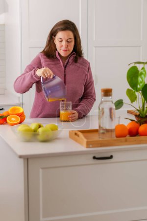Téléchargez les photos : Close-up of an attractive young brunette girl pouring orange juice with a pitcher into a drinking glass - en image libre de droit