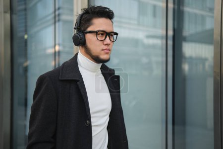 Foto de An Asian guy businessman in a headphones walks past office building. - Imagen libre de derechos