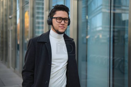 Photo for An Asian guy businessman in a headphones walks past office building. - Royalty Free Image