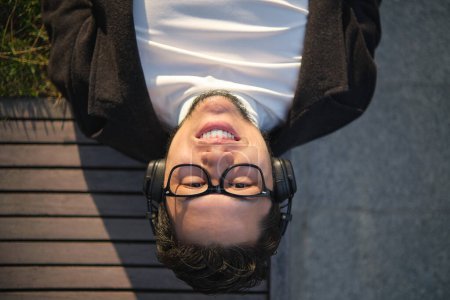 Photo for Asian guy in glasses and headphones, resting lying on a wooden bench - Royalty Free Image