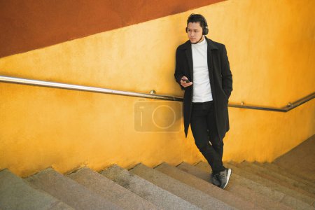 Foto de An Asian guy in a headphones climbs the stairs behind him yellow wall - Imagen libre de derechos