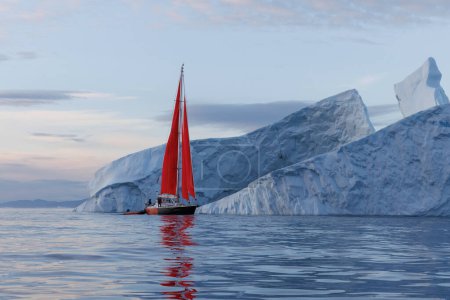 sailboat with red sails sailing between huge icebergs