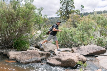 Foto de Hombre en camiseta, corto, con la mochila saltando sobre un río en un n - Imagen libre de derechos