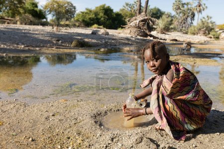 Foto de Young Himba filling a water bottle in Kunene river, Epupa, Namibia - Imagen libre de derechos