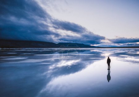 Téléchargez les photos : Lone figure stands on frozen lake with reflection and storm clouds - en image libre de droit