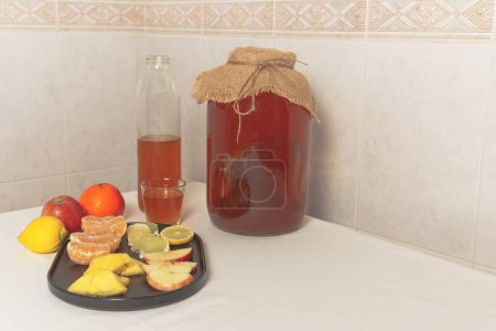 Foto de Large glass bottle with fermented kombucha tea on the white kitchen table and assorted fruit for taste - Imagen libre de derechos