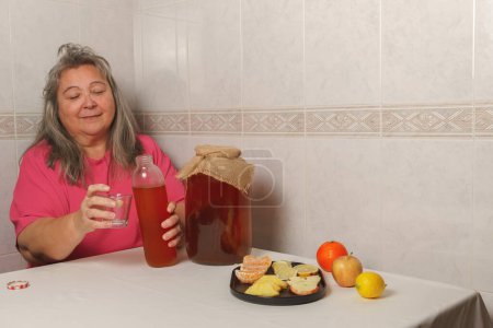 Téléchargez les photos : Older white-haired woman filling a glass of kombucha tea, with assorted fruits and large bottle of fermented tea. - en image libre de droit