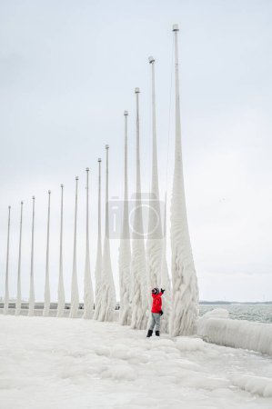 Photo for Child in red coat touching icy flagpoles on frozen pier in winter. - Royalty Free Image
