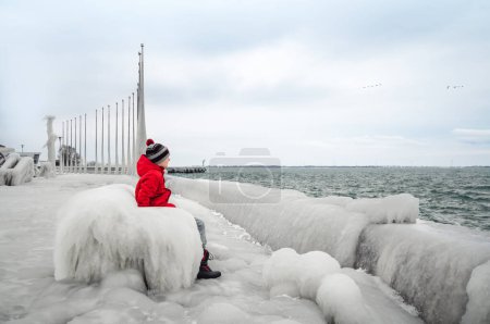 Photo for Child in red coat sitting on icy bench on frozen pier in winter. - Royalty Free Image