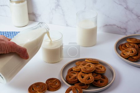 Téléchargez les photos : A man's hand pours Milk in a glass. Glass bottle with milk on a white table, cookies in a plate - en image libre de droit