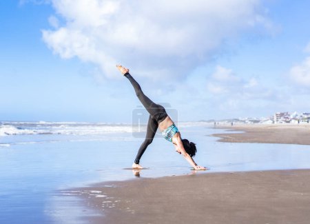 Téléchargez les photos : A woman practicing yoga on the water at the beach, doing Three Legged Downward-Facing Dog (Eka Pada Adho Mukha Shvanasana) pose. - en image libre de droit
