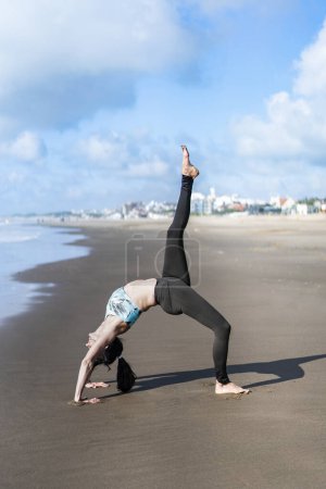 Foto de A woman practicing yoga on the water at the beach, doing One Legged Wheel (Eka Pada Urdhva Dhanurasana) pose. - Imagen libre de derechos