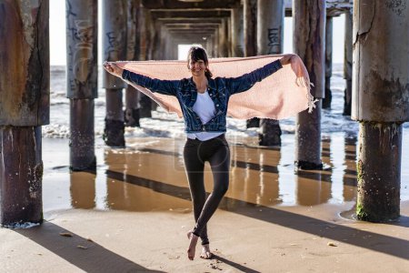 Foto de Portrait of a happy woman under a pier holding a scarf on the wind while looking at camera - Imagen libre de derechos