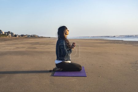 Téléchargez les photos : Side view of a woman sitting on the sand while meditating at the beach at sunrise - en image libre de droit
