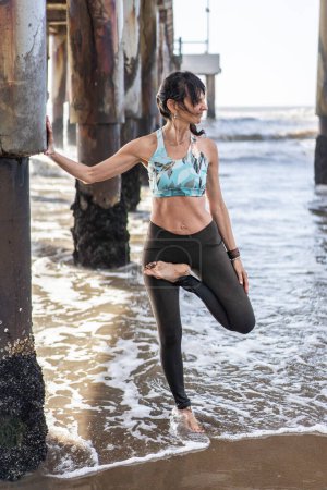 Téléchargez les photos : A woman doing stretching exercises under a pier at the beach - en image libre de droit