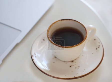 Téléchargez les photos : A mug with fragrant black coffee stands on a white table. Behind is a laptop - en image libre de droit