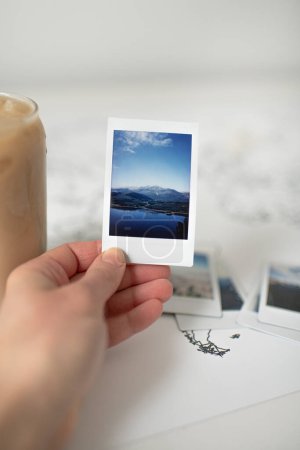 Téléchargez les photos : Polaroïd main tenant la photo de montagne sur la carte du monde avec du café glacé - en image libre de droit