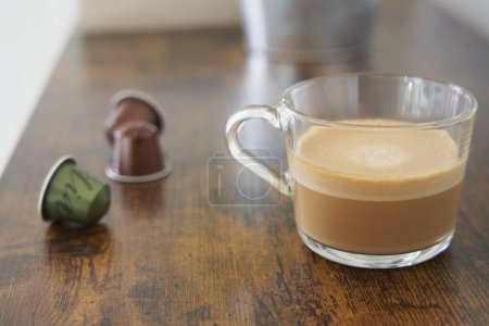 Téléchargez les photos : Cup of foamy coffee with milk next to some capsules on table - en image libre de droit