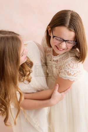 Photo for Sisters in lace dresses hugging and laughing against pink backdrop - Royalty Free Image