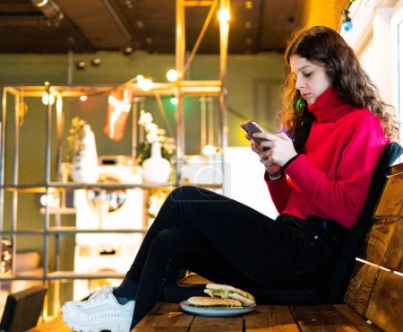 Téléchargez les photos : Femme assise dans un hôtel regardant le mobile avec panini dans un plat - en image libre de droit