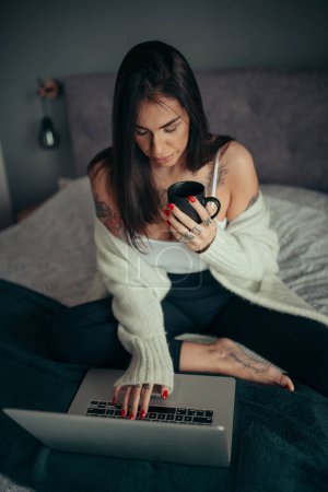Photo for Tattooed woman with cup of coffee using laptop on the bed. - Royalty Free Image