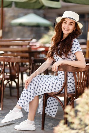 Téléchargez les photos : Jeune femme heureuse dans une robe et un chapeau est assis à une table dans un café - en image libre de droit
