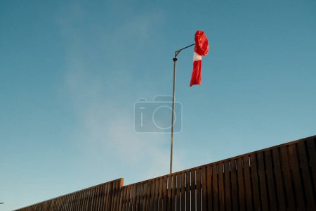 Photo for From below of striped red and white windsock hanging on pole against cloudy blue sky near wooden fence - Royalty Free Image