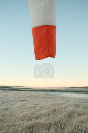 Photo for Red and white striped wind sleeve hanging against cloudless sundown blue sky over dry grassy field in peaceful weather - Royalty Free Image