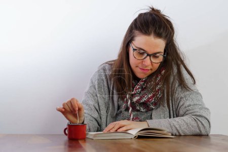 Téléchargez les photos : Young brunette girl with glasses reading a book while drinking coffee - en image libre de droit
