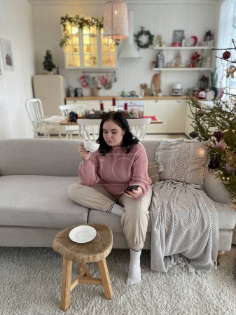 Téléchargez les photos : Jeune fille boit du café et tenant smartphone assis sur un canapé - en image libre de droit