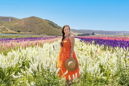 Foto de Mujer en vestido naranja mirando a la cámara, de pie en el campo de flores - Imagen libre de derechos