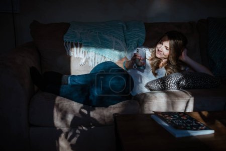 Photo for Woman lounging on sofa watching mobile phone in home - Royalty Free Image