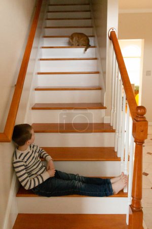 Foto de Gato mascota naranja mira por las escaleras al niño sentado con las piernas extendidas - Imagen libre de derechos