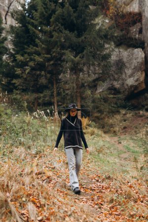Photo for Woman in a jacket and hat walks through the woods near the mountains - Royalty Free Image