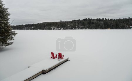 Téléchargez les photos : Red adirondack chairs on end of snowy dock on a lake in winter. - en image libre de droit