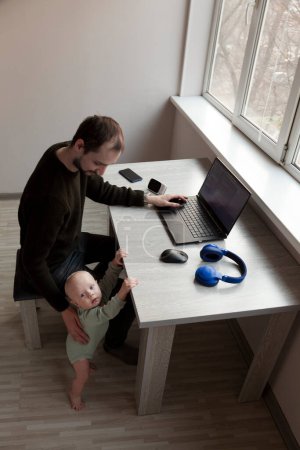 Téléchargez les photos : Jeune homme travaillant sur un ordinateur portable à la maison avec un enfant - en image libre de droit