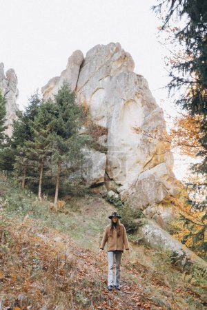 Foto de Mujer con chaqueta y sombrero camina por el bosque cerca de las montañas - Imagen libre de derechos