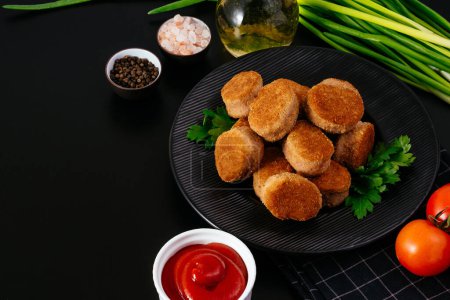 Téléchargez les photos : Fried chicken nuggets on a black plate, ketchup in a saucepan, vegetables on a background nearby. - en image libre de droit