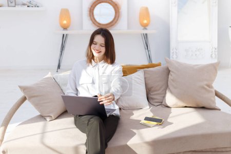 Téléchargez les photos : Jeune femme souriante dans des vêtements de style d'affaires travaille dans un ordinateur portable à la maison - en image libre de droit