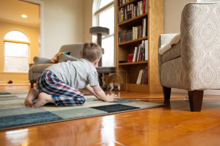 Photo for Playful Boy Crouches Down on Living Room Floor as Cat Walks Away - Royalty Free Image