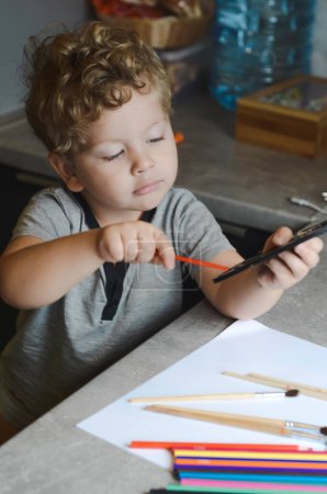 Téléchargez les photos : Blanc bouclé garçon dessine avec des peintures et des crayons - en image libre de droit