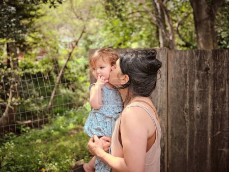 Foto de Mamá besos niño en la mejilla en vallado en el patio trasero lleno de árboles - Imagen libre de derechos