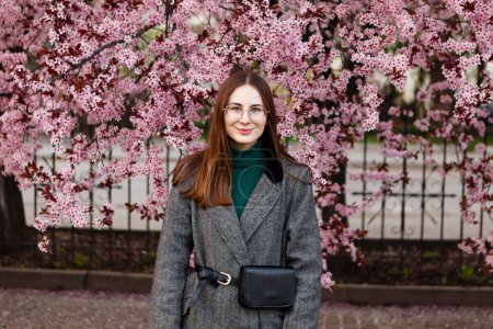 Téléchargez les photos : Jeune femme en manteau de laine marche parmi les fleurs de cerisier au printemps - en image libre de droit