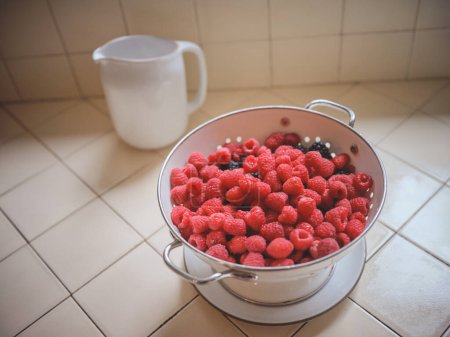 Foto de Raspberries in a colander with water pitcher on kitchen counter - Imagen libre de derechos